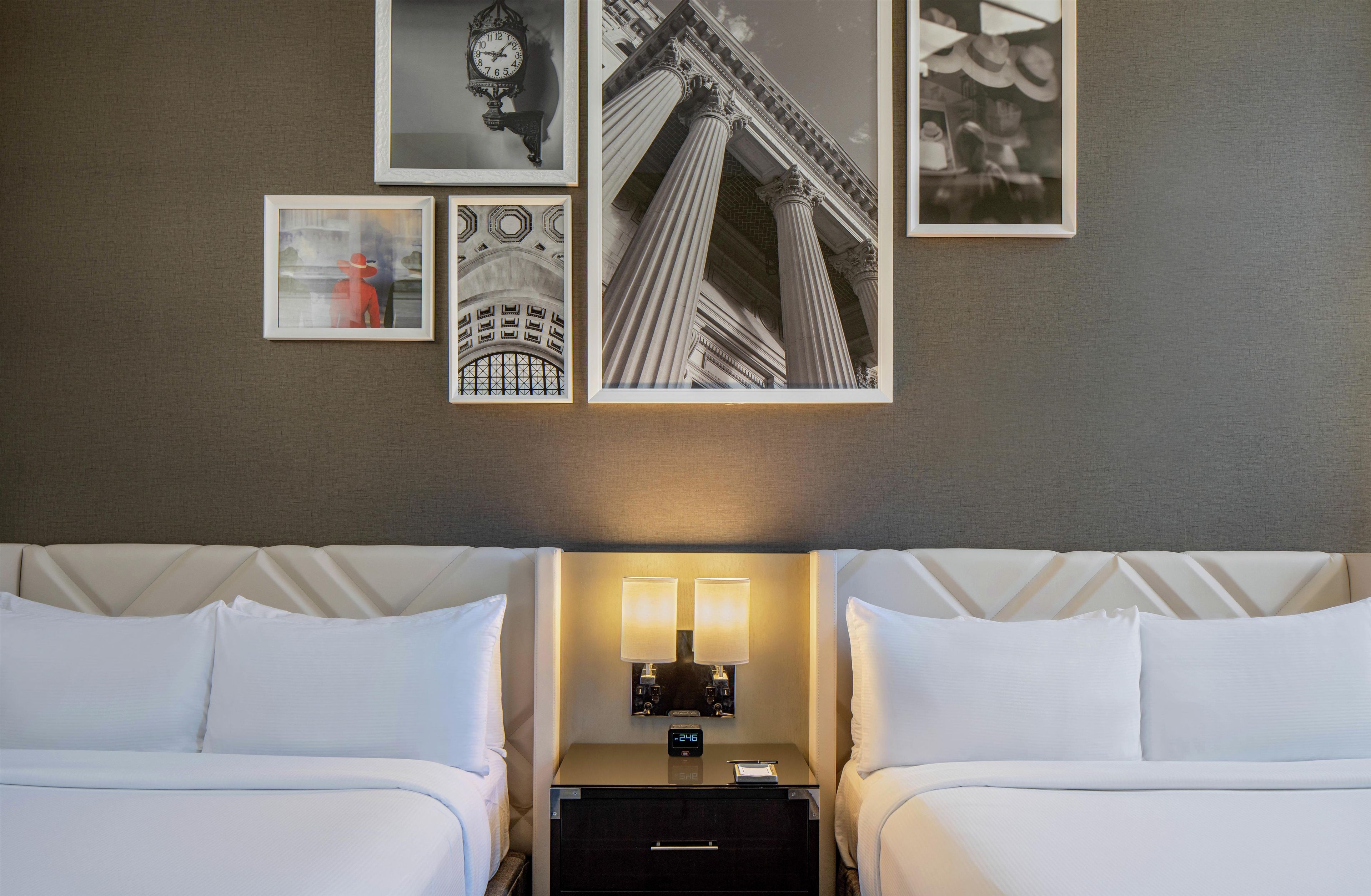 A hotel room with two beds, a nightstand with a lamp, and wall art including a clock and architectural images.