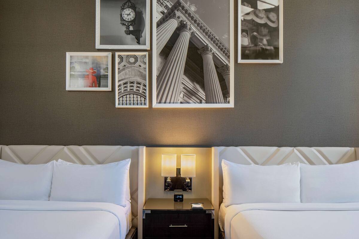 A hotel room with two beds, a nightstand with a clock, and framed pictures on the wall.