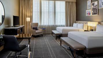 A hotel room with a desk, two beds, a chair, a small table, and a large window with curtains.
