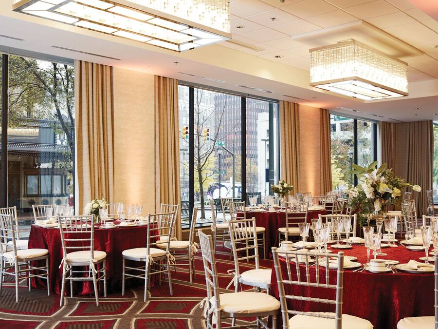 Uma sala de banquetes com várias mesas redondas preparadas para refeições, grandes janelas com vista para a cidade e um candelabro pendurado no teto.