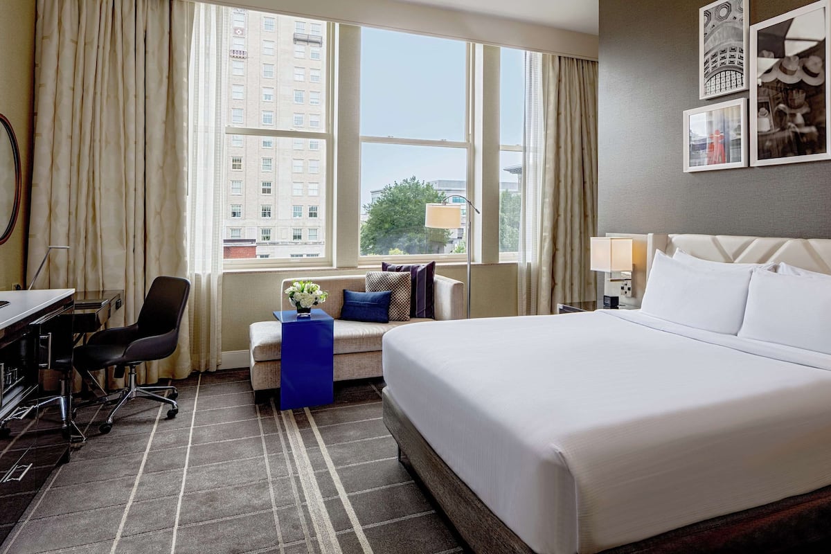 A hotel room with a large bed, a desk with a chair, a small table with a vase, and a window with curtains.