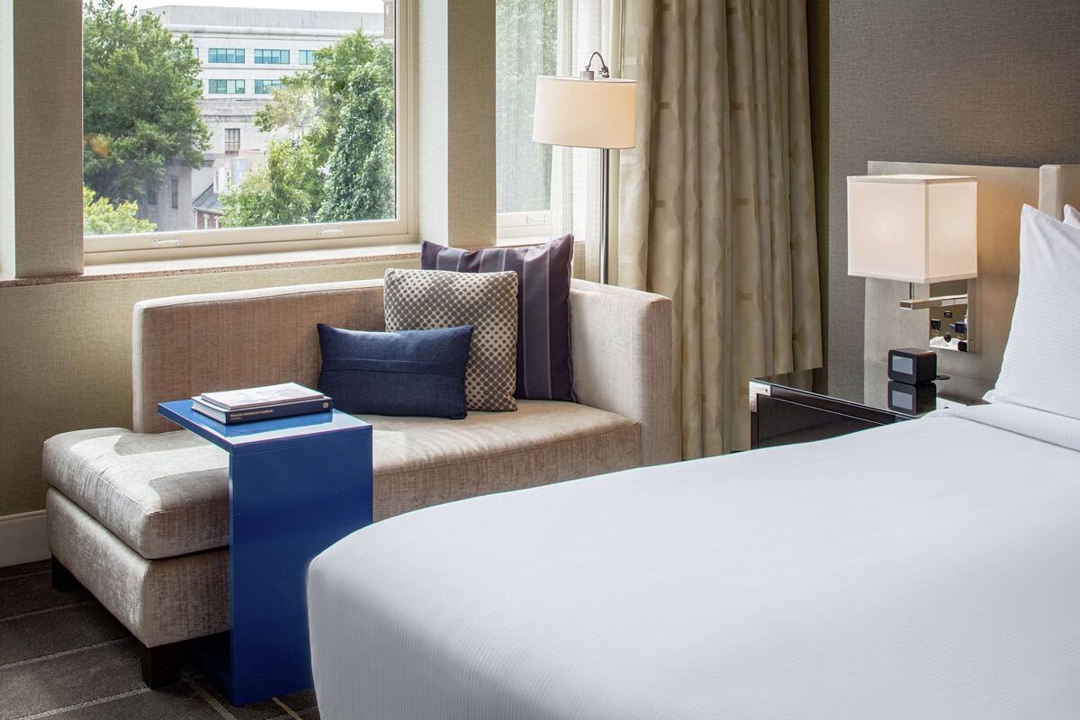 A hotel room with a bed, a sofa, a small table with a book, a lamp, and a view of a building through the window.