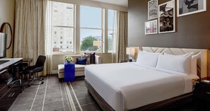 Habitación de hotel con una cama grande, un escritorio y una silla. Se ve un ventanal con vistas a edificios y árboles.