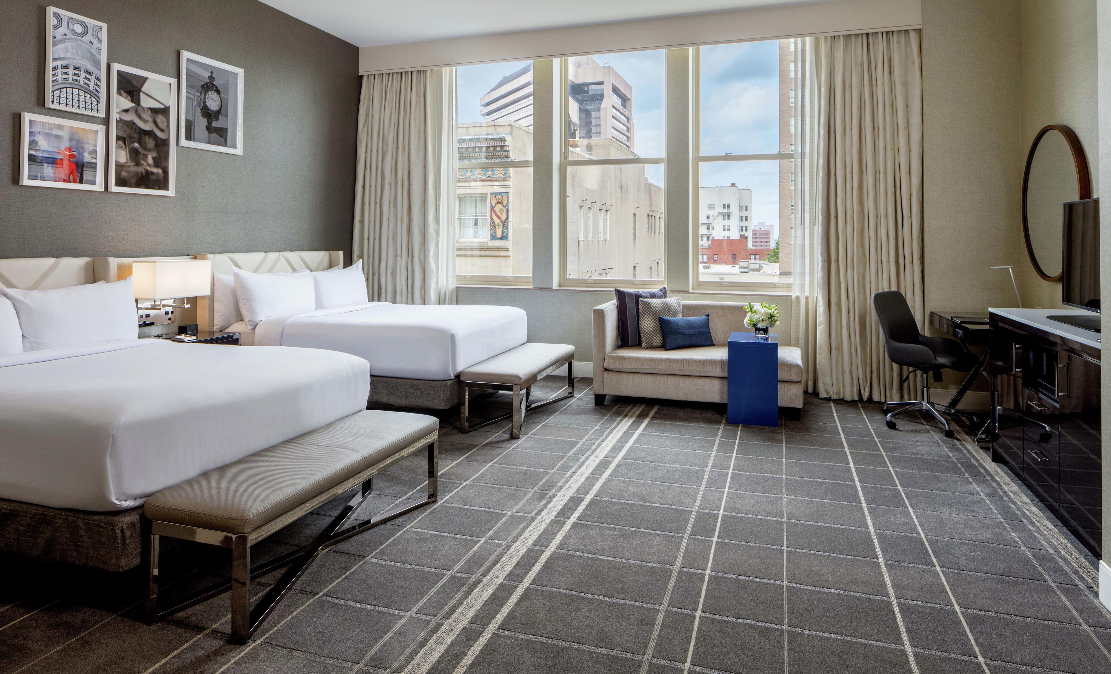 A hotel room with two beds, a sofa, a desk, and a large window with a city view.