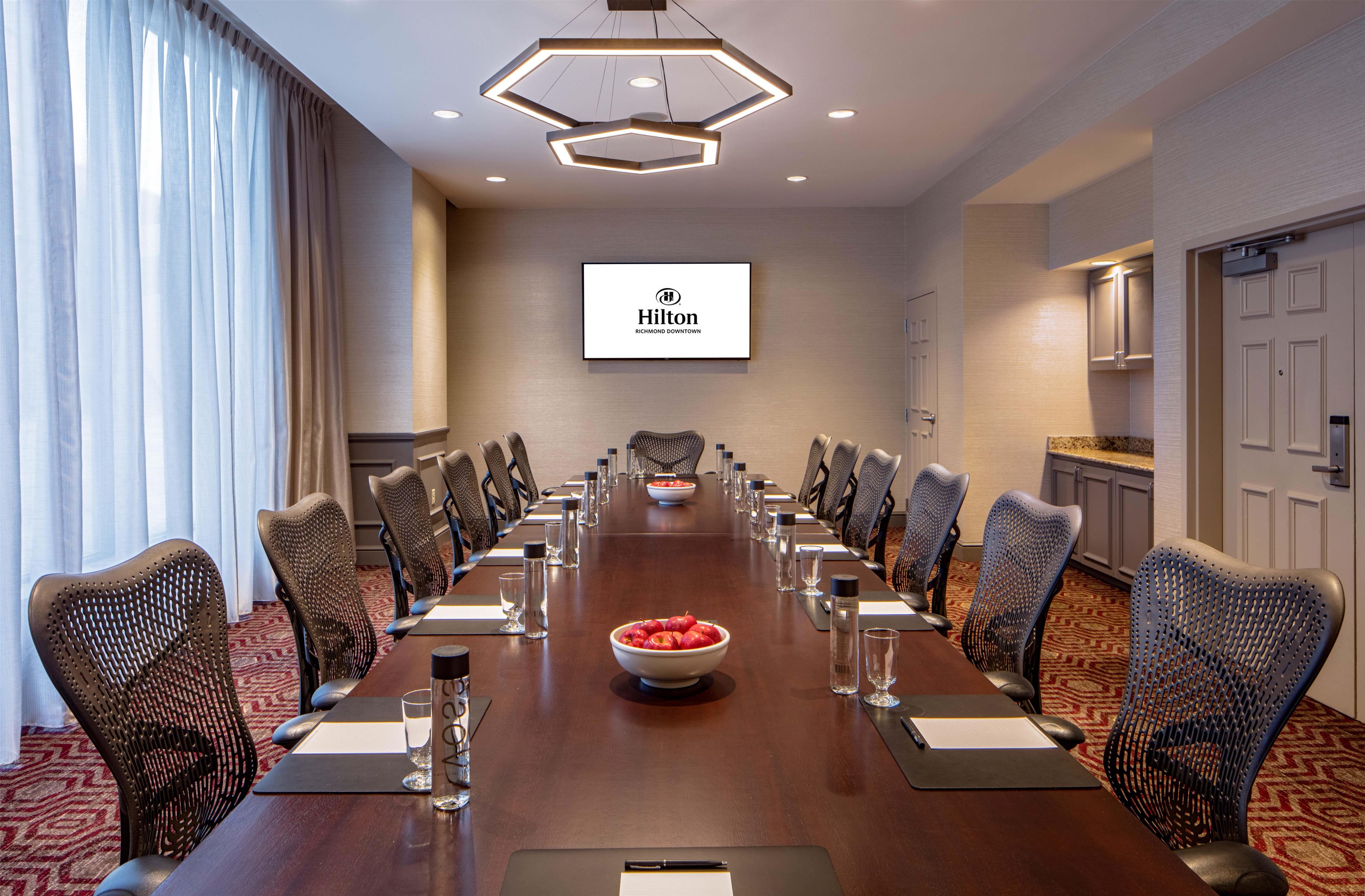 Un salón de conferencias con una mesa larga, sillas, un frutero y el logotipo de Hilton en la pared.