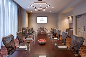 Una sala conferenze con un lungo tavolo, sedie, una ciotola di frutta e il logo Hilton sulla parete.