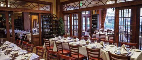 A restaurant with multiple dining tables set for guests, large windows offering a view, and a chandelier hanging from the ceiling.