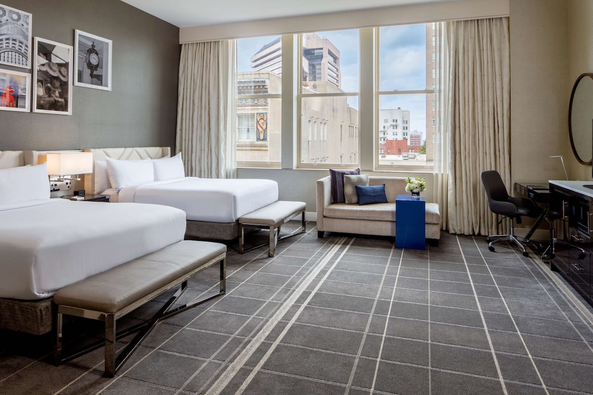 A hotel room with two beds, a sofa, a desk, and a large window with a city view.
