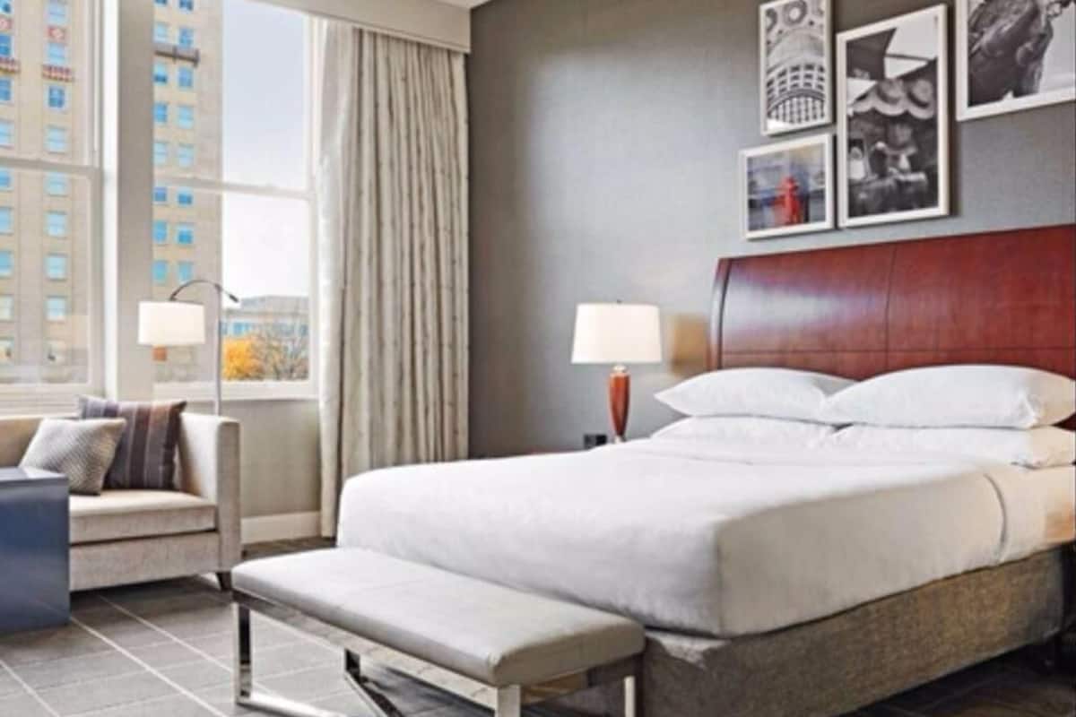 A hotel room with a bed, a bench, a desk, a lamp, and a window with curtains.