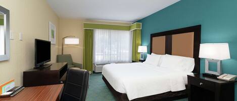 A hotel room with a bed, a desk with a computer, a TV, and a nightstand with a lamp.