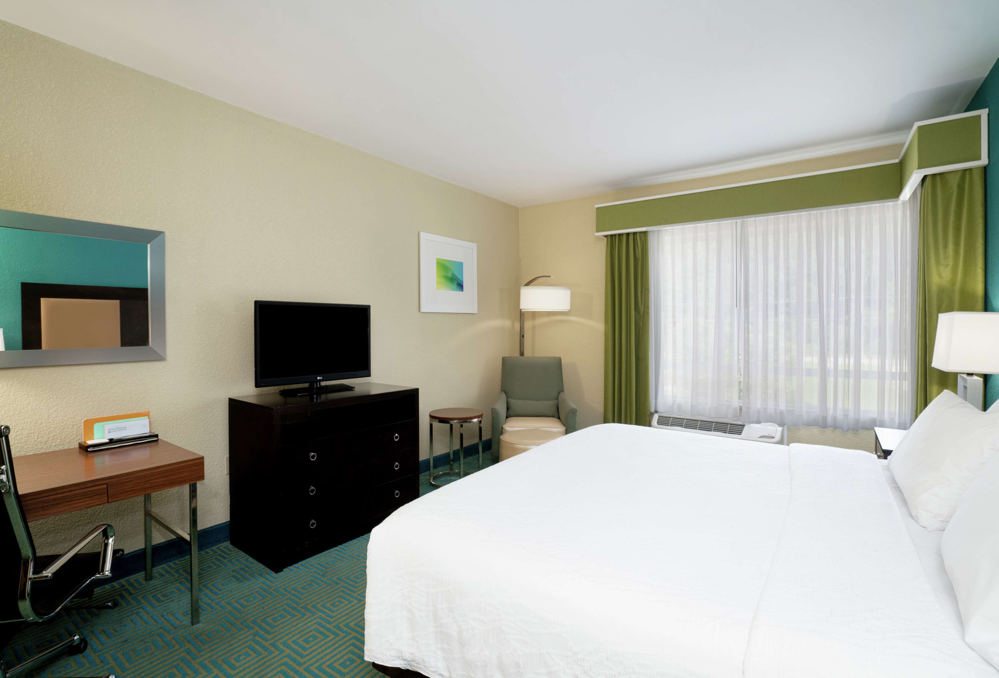 A hotel room with a large bed, a desk with a computer, a television, a chair, and a window with curtains.