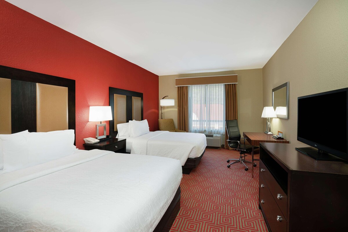 A hotel room with two beds, a desk, a television, and a window with curtains.
