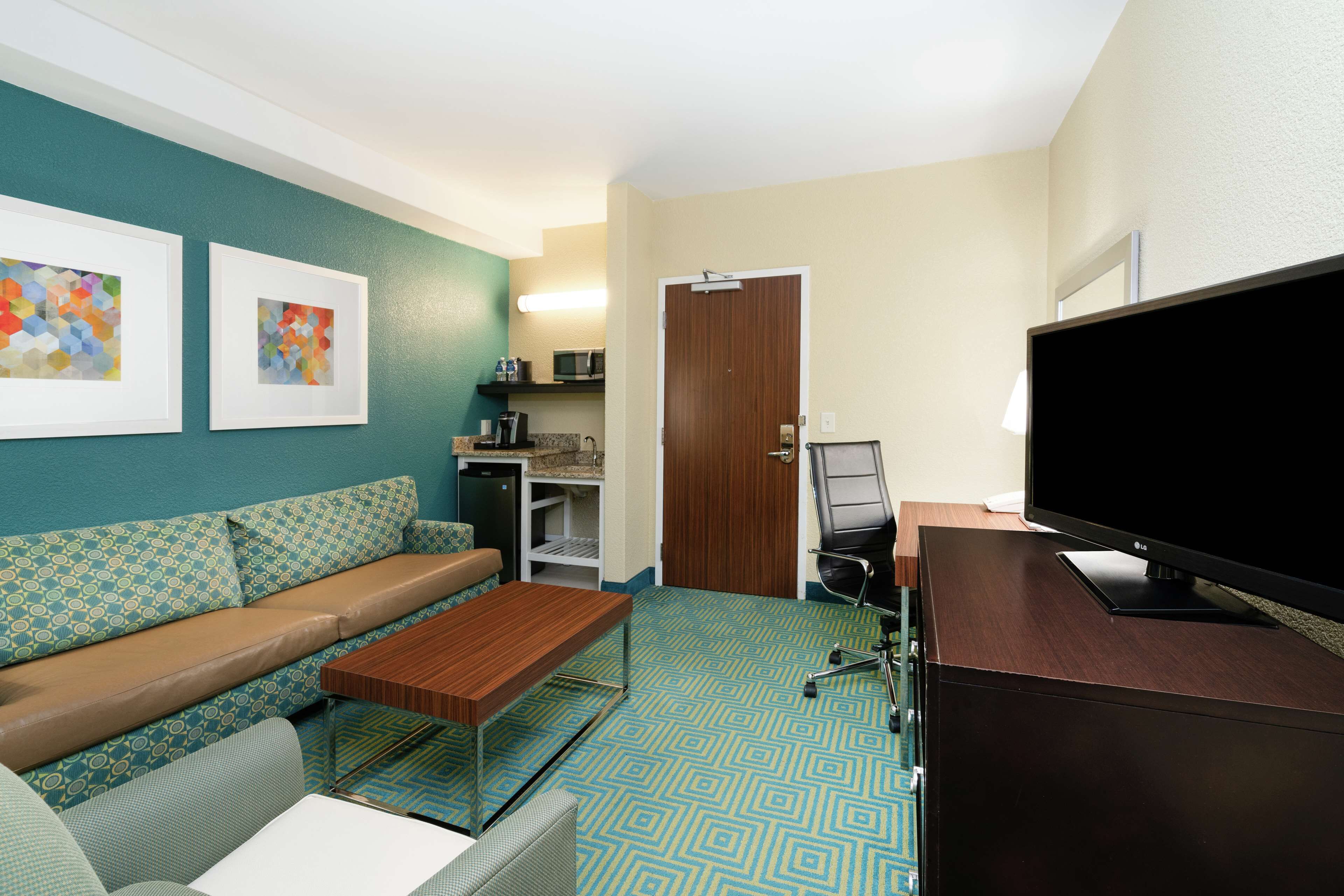 A hotel room with a sofa, a coffee table, a TV, a desk with a computer, and artwork on the wall.