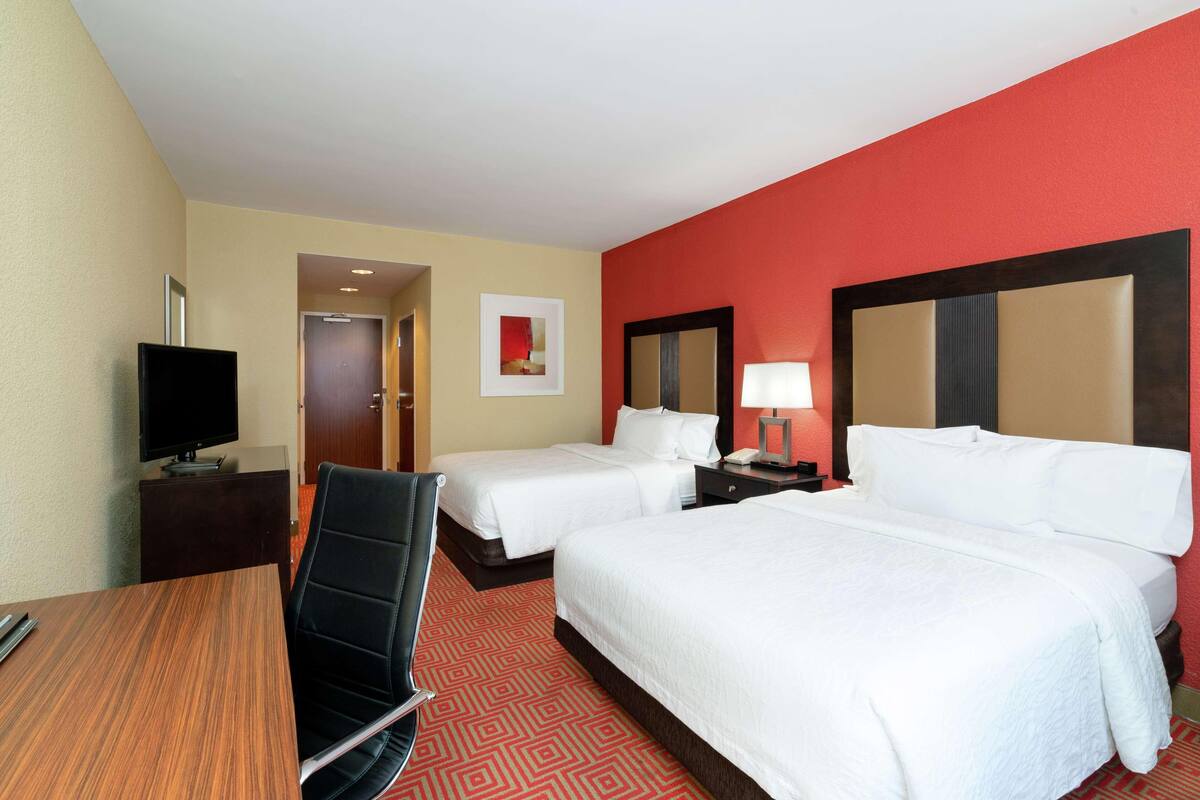 A hotel room with two beds, a desk with a chair, a television, and a red carpet.