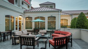 An outdoor patio area with seating arrangements, a fire pit, and a view of the Hilton Garden Inn building.