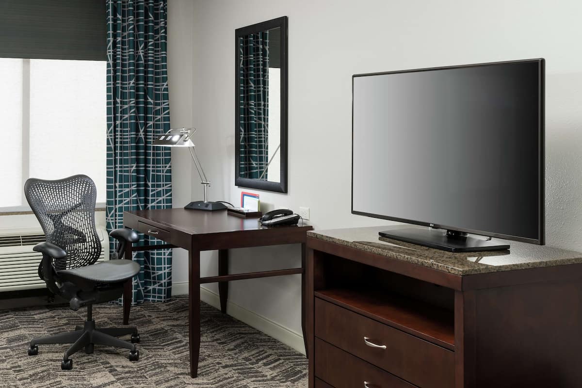 A hotel room with a desk, chair, television, and a large window with curtains.