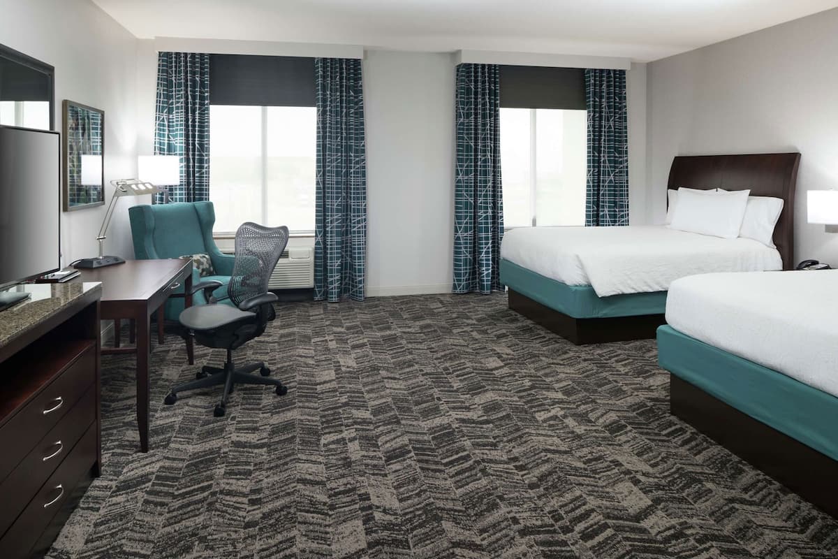 A hotel room with a desk, office chair, two beds, and a large window with curtains.