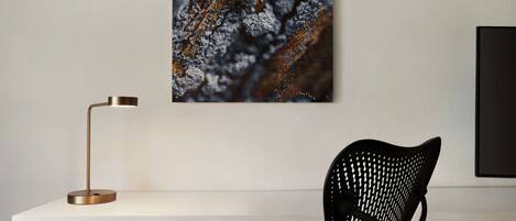 A modern desk with a metallic floor lamp, a black woven chair, and a framed artwork on the wall.