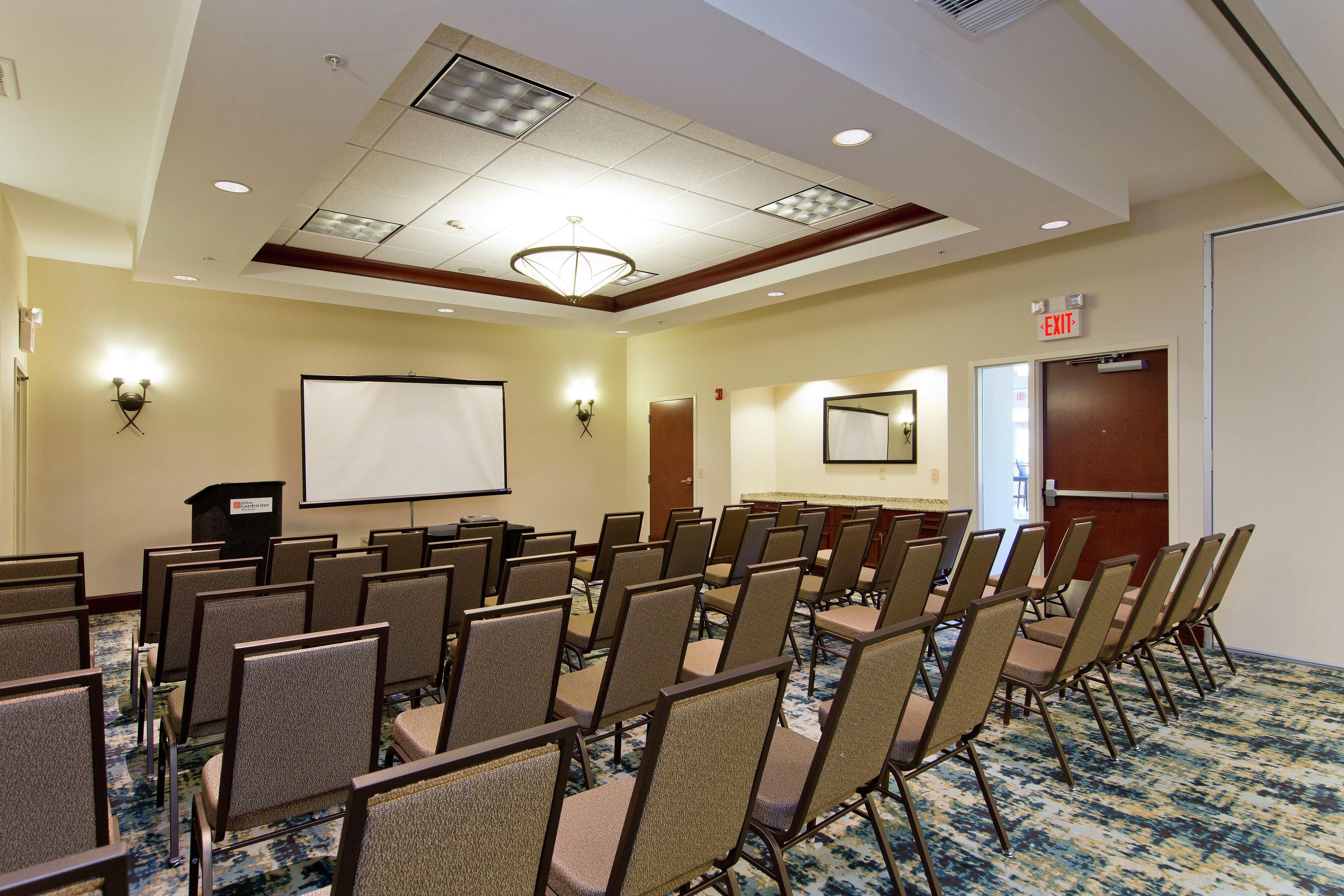 A conference room with rows of chairs, a projector screen, and an exit door.