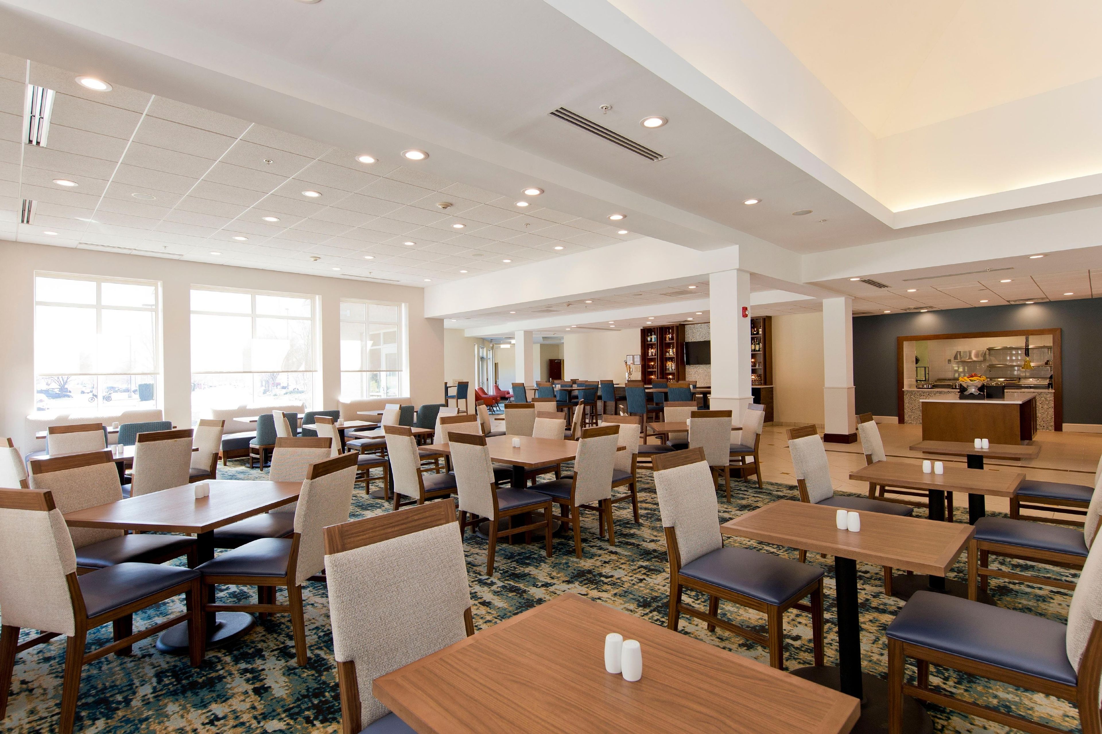 A spacious hotel dining area with multiple tables and chairs, large windows, and a kitchen area visible in the background.