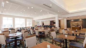 A spacious hotel dining area with multiple tables and chairs, large windows, and a kitchen area visible in the background.
