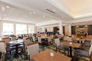 A spacious hotel dining area with multiple tables and chairs, large windows, and a kitchen area visible in the background. - Hilton Garden Inn Winchester (Winchester)