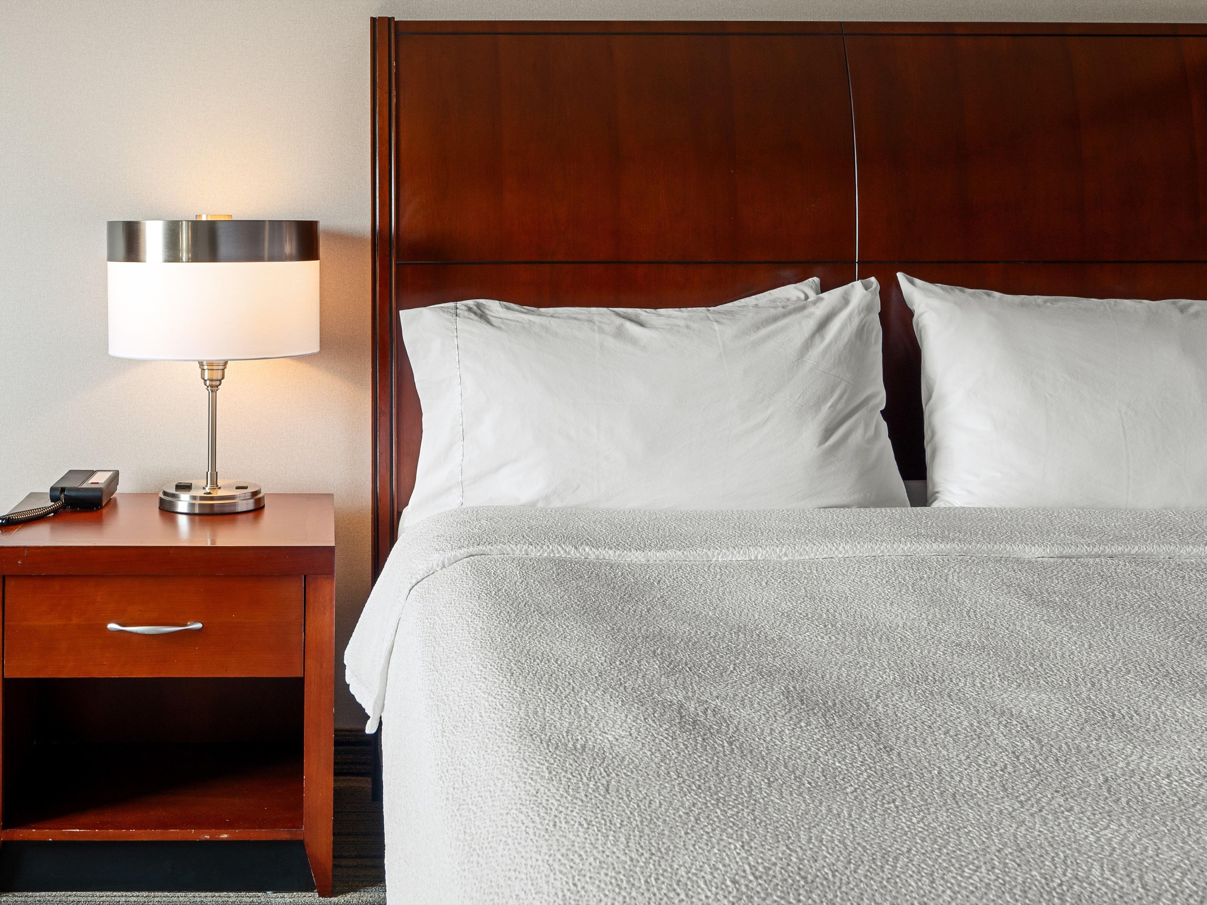 A hotel room with a wooden headboard, a bedside table with a lamp, and a neatly made bed with white linens.