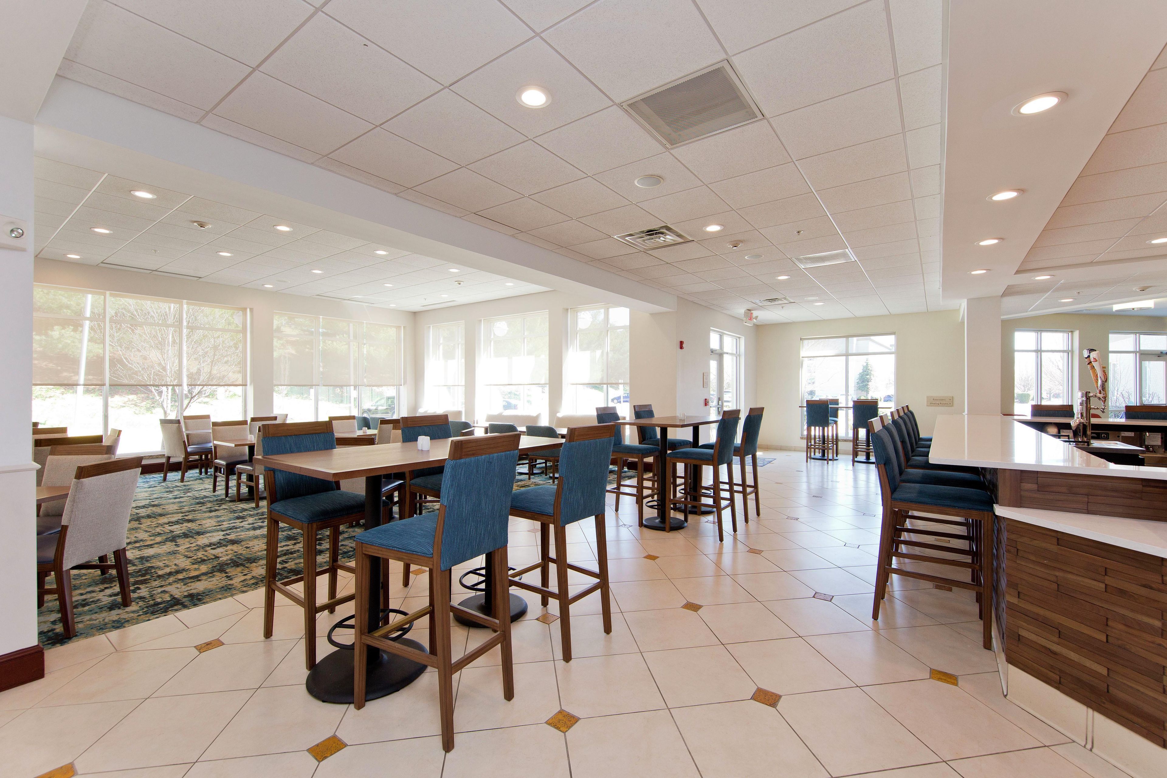 A spacious dining area with multiple tables and chairs, large windows allowing natural light, and a clean, tiled floor.