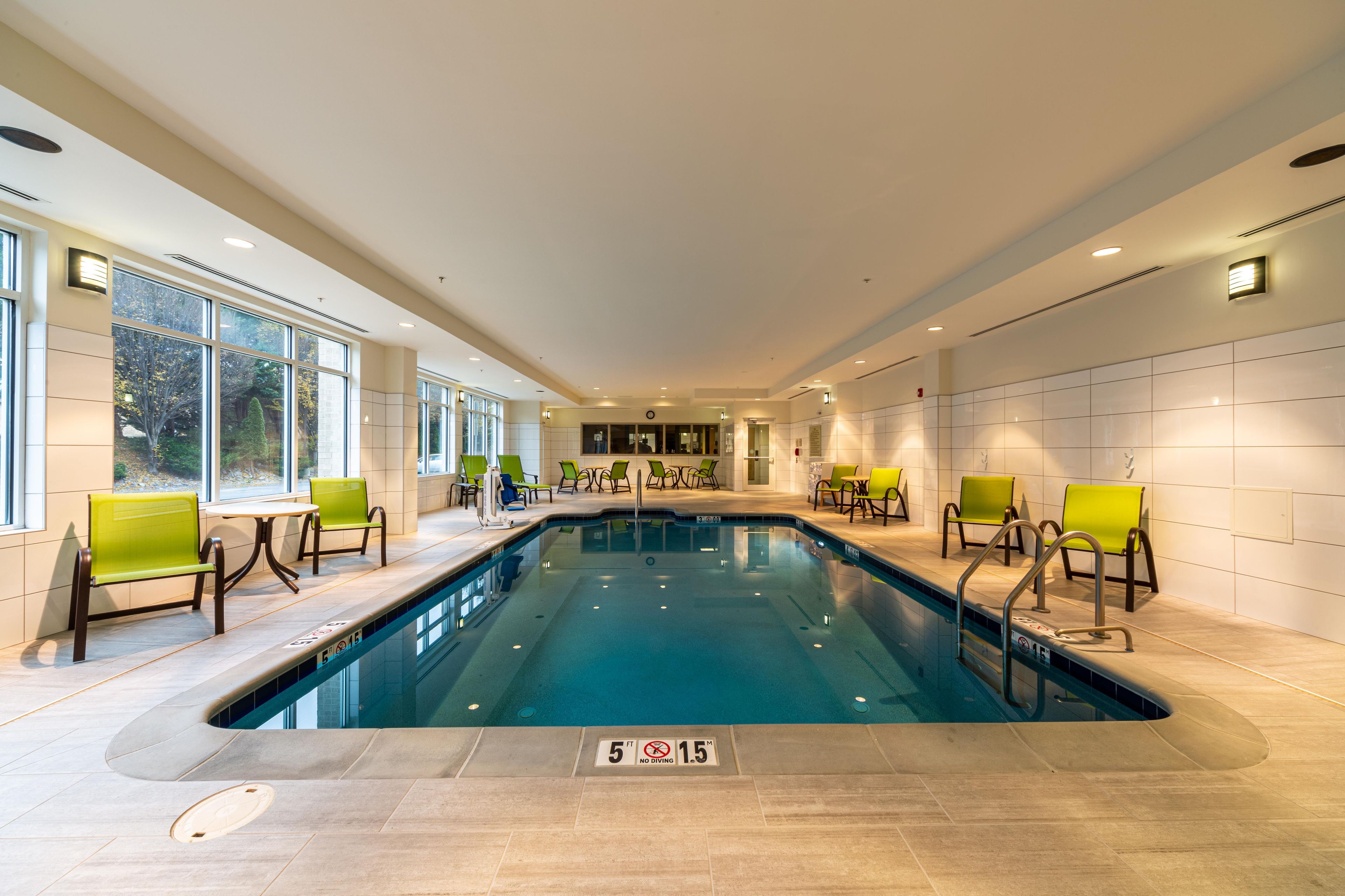 An indoor swimming pool with clear water, surrounded by green chairs and tables, and large windows on one side.
