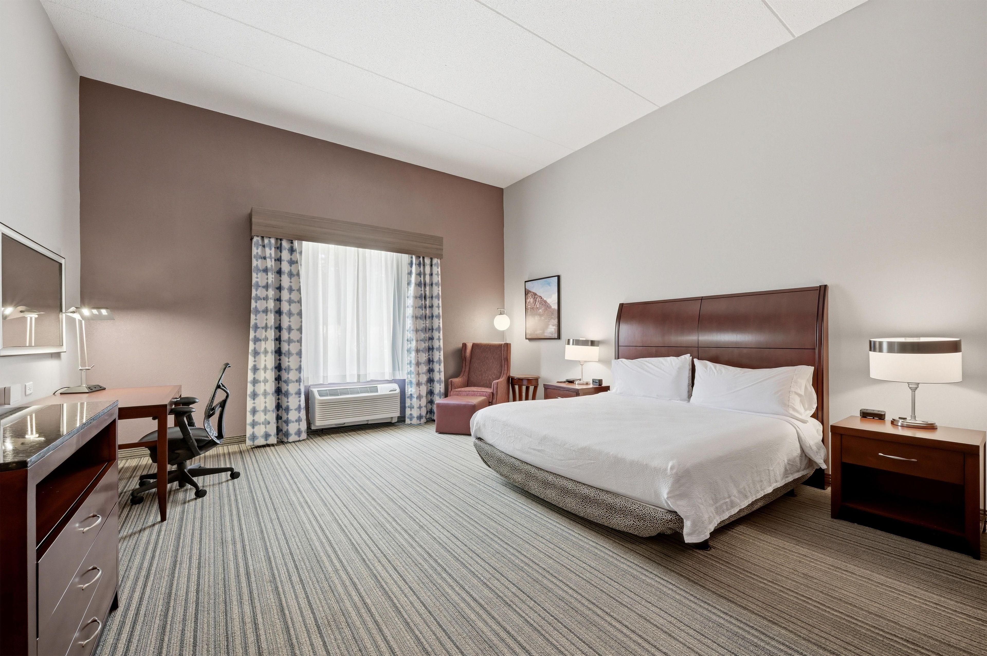 Une chambre d’hôtel avec un grand lit, un bureau avec une chaise, une petite table, une chaise, une lampe et un tableau au mur.