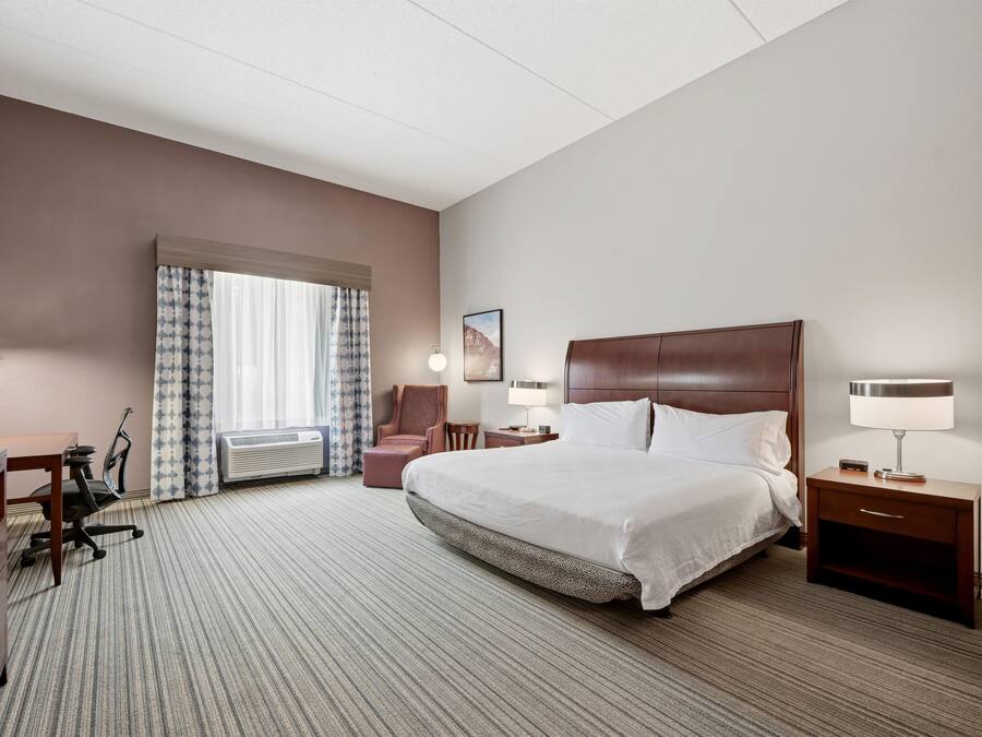 A hotel room with a large bed, a desk with a chair, a small table, a chair, a lamp, and a picture on the wall.