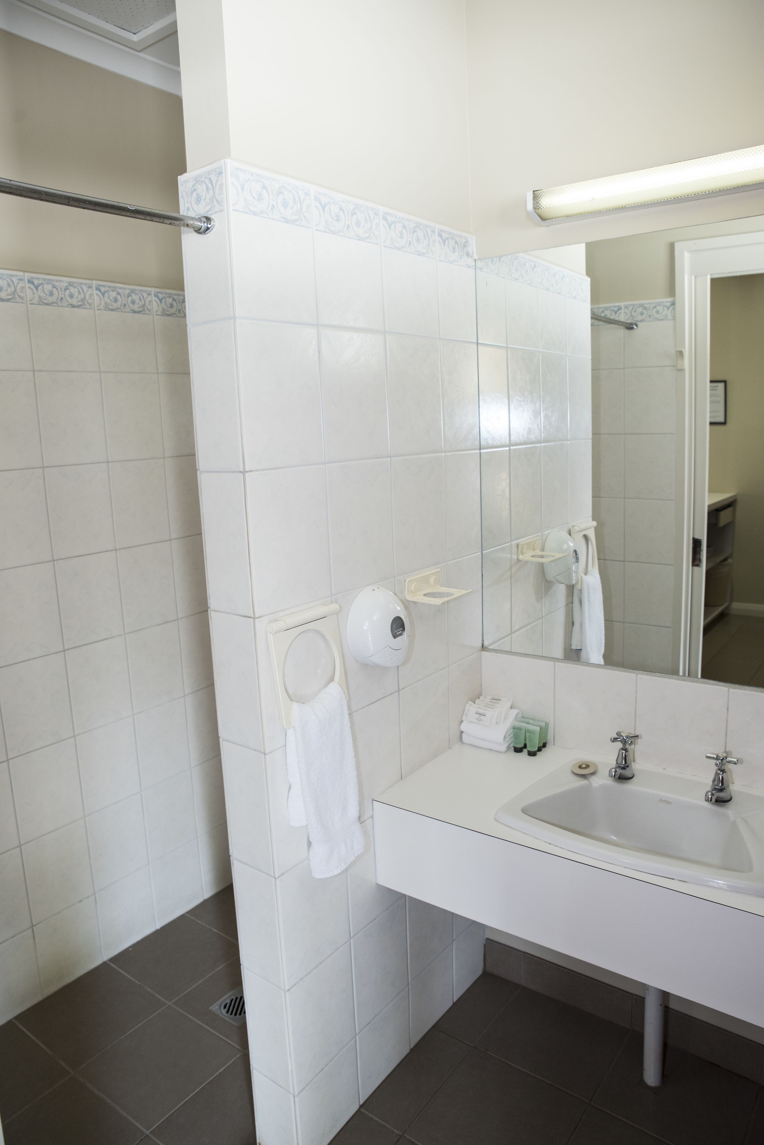 Un baño con azulejos blancos, un lavamanos, un espejo, un perchero para toallas y un dispensador de papel higiénico.