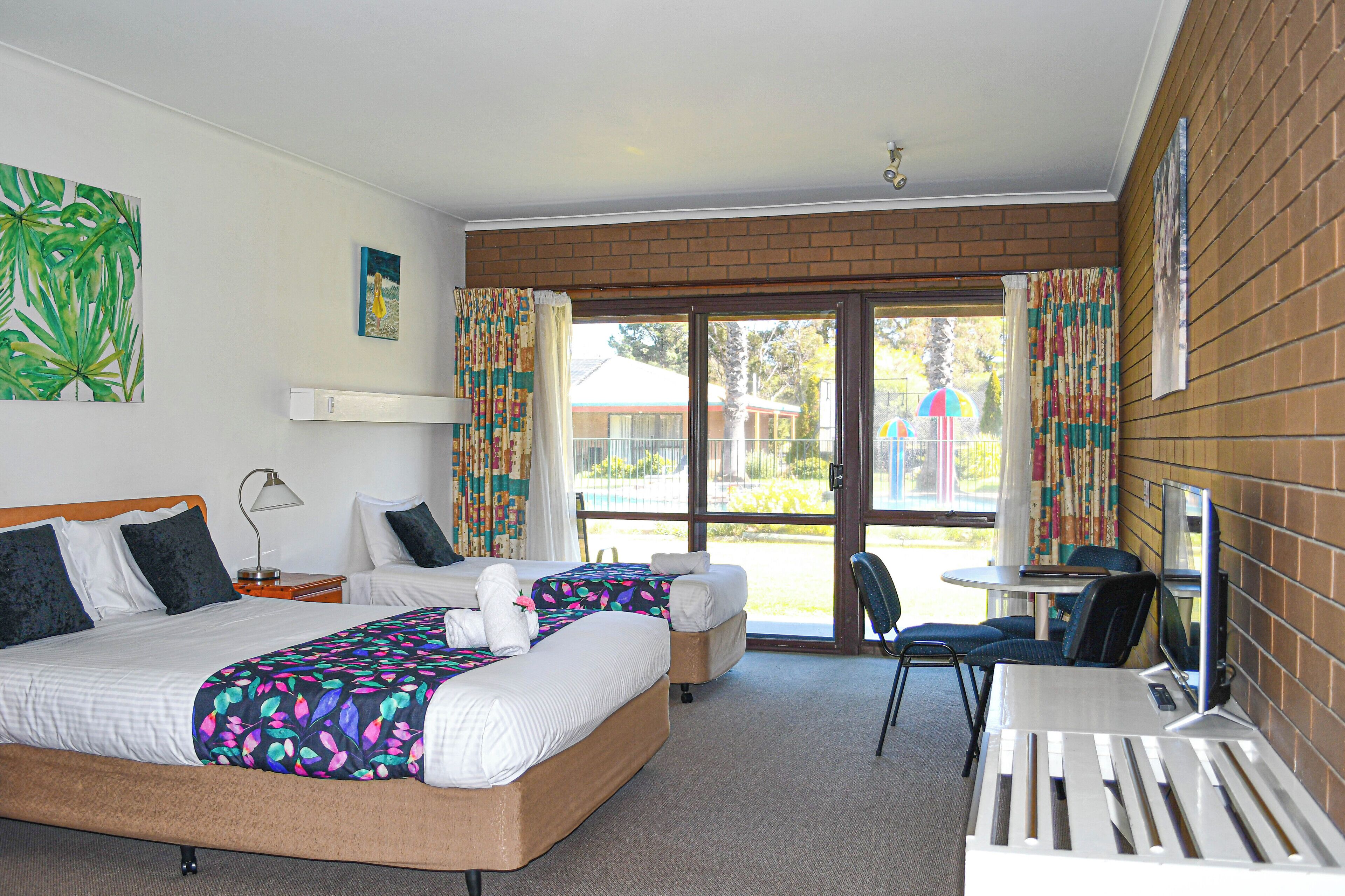 A hotel room with two beds, a desk, and a chair. There is a window with curtains and a view of the outside.