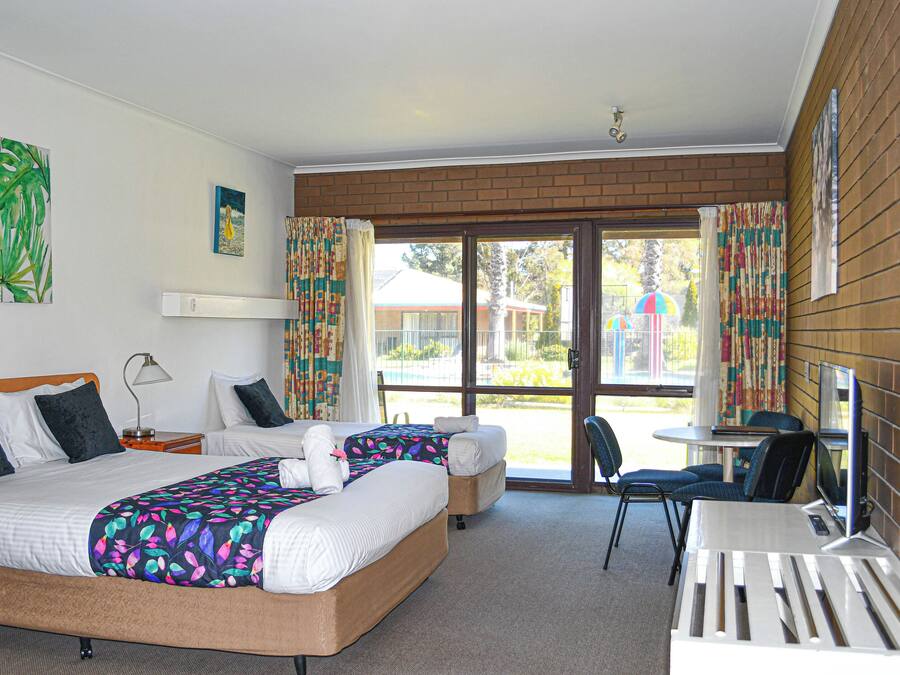 A hotel room with two beds, a desk, and a chair. There is a window with curtains and a view of the outside.