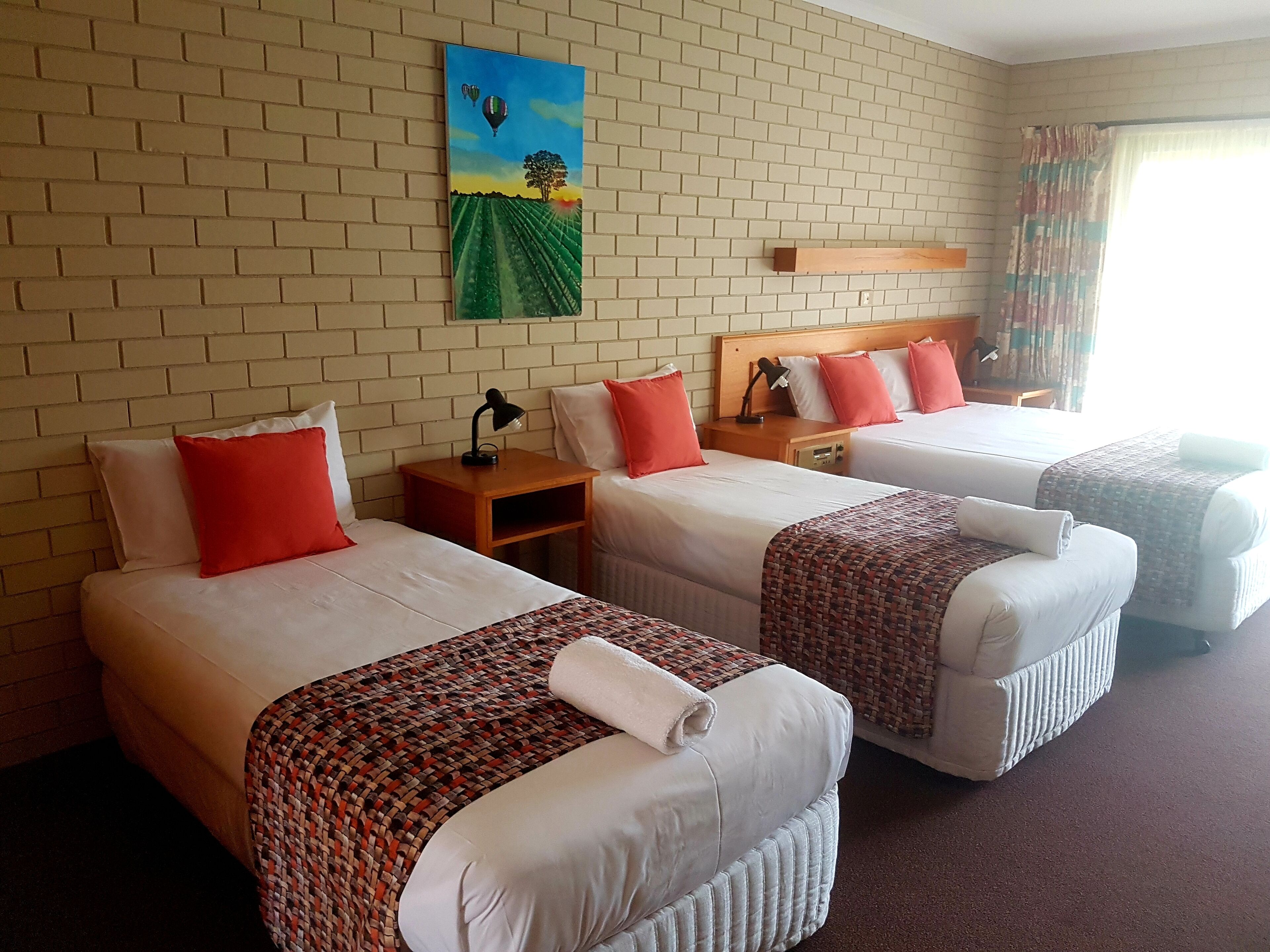 A hotel room with two single beds, a large window with curtains, a brick wall, and a painting of a landscape.