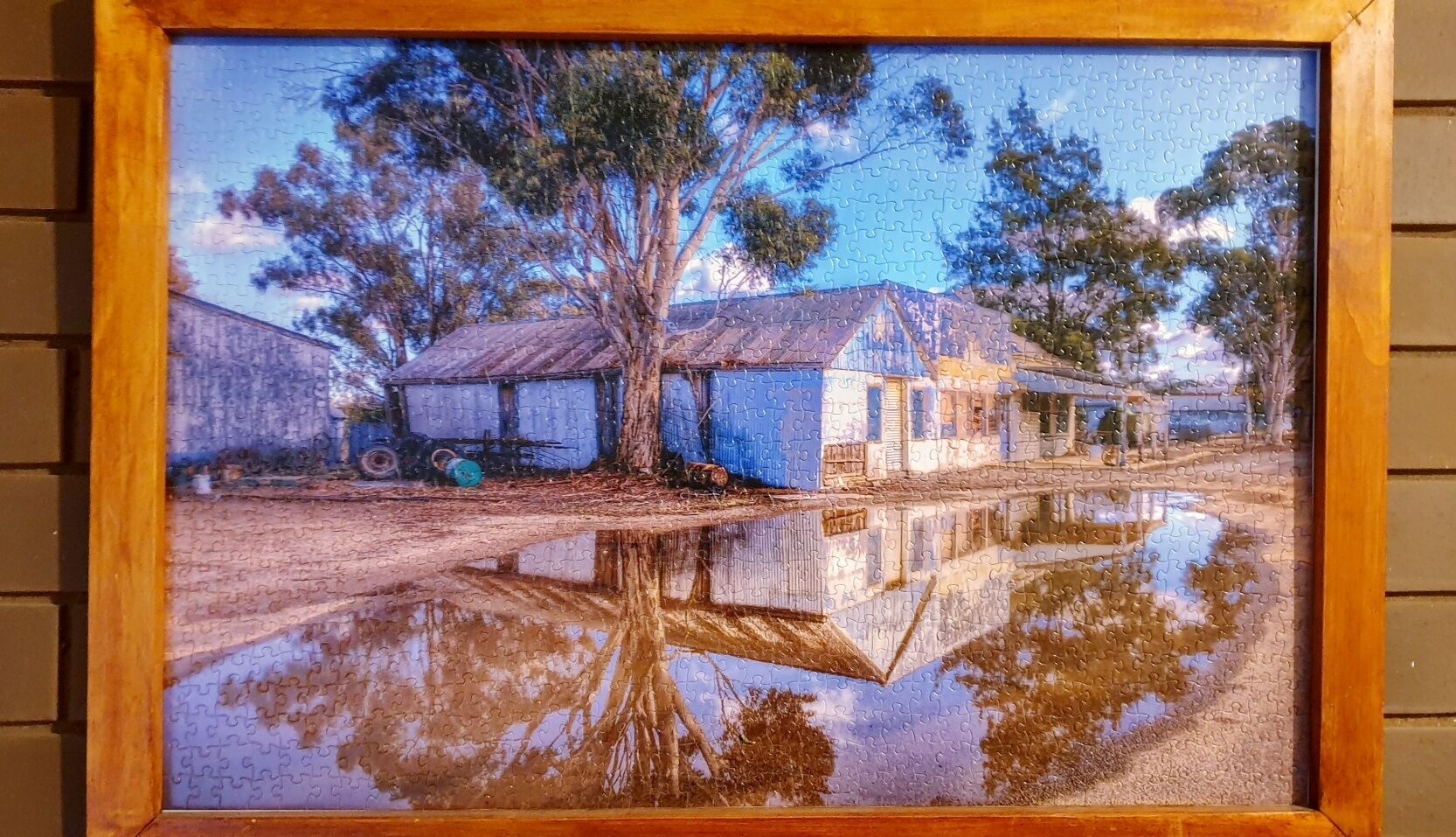 A framed painting of a rural scene with houses and trees reflected in a puddle.