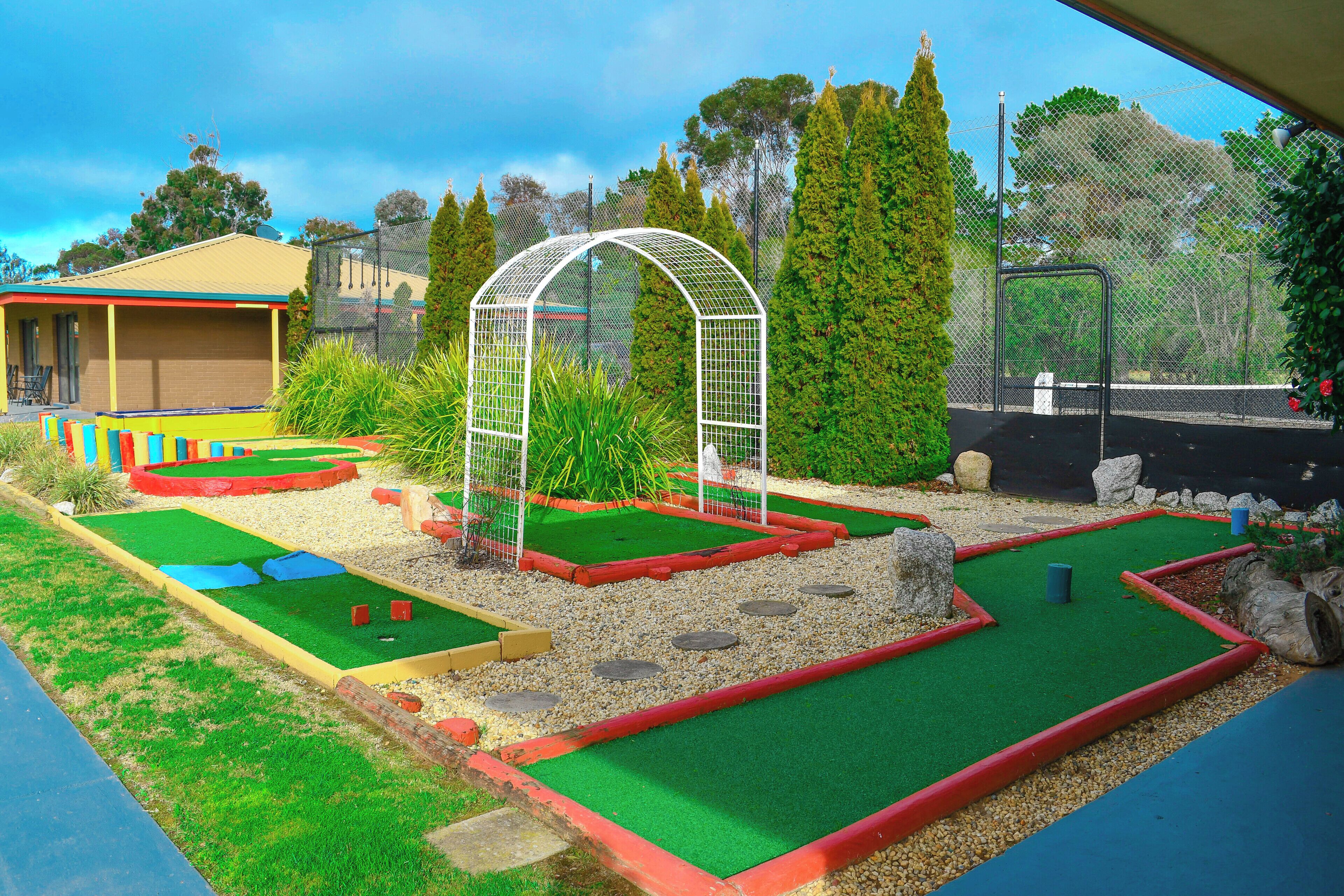 A miniature golf course with multiple green putting areas, a white arch structure, and surrounding greenery.