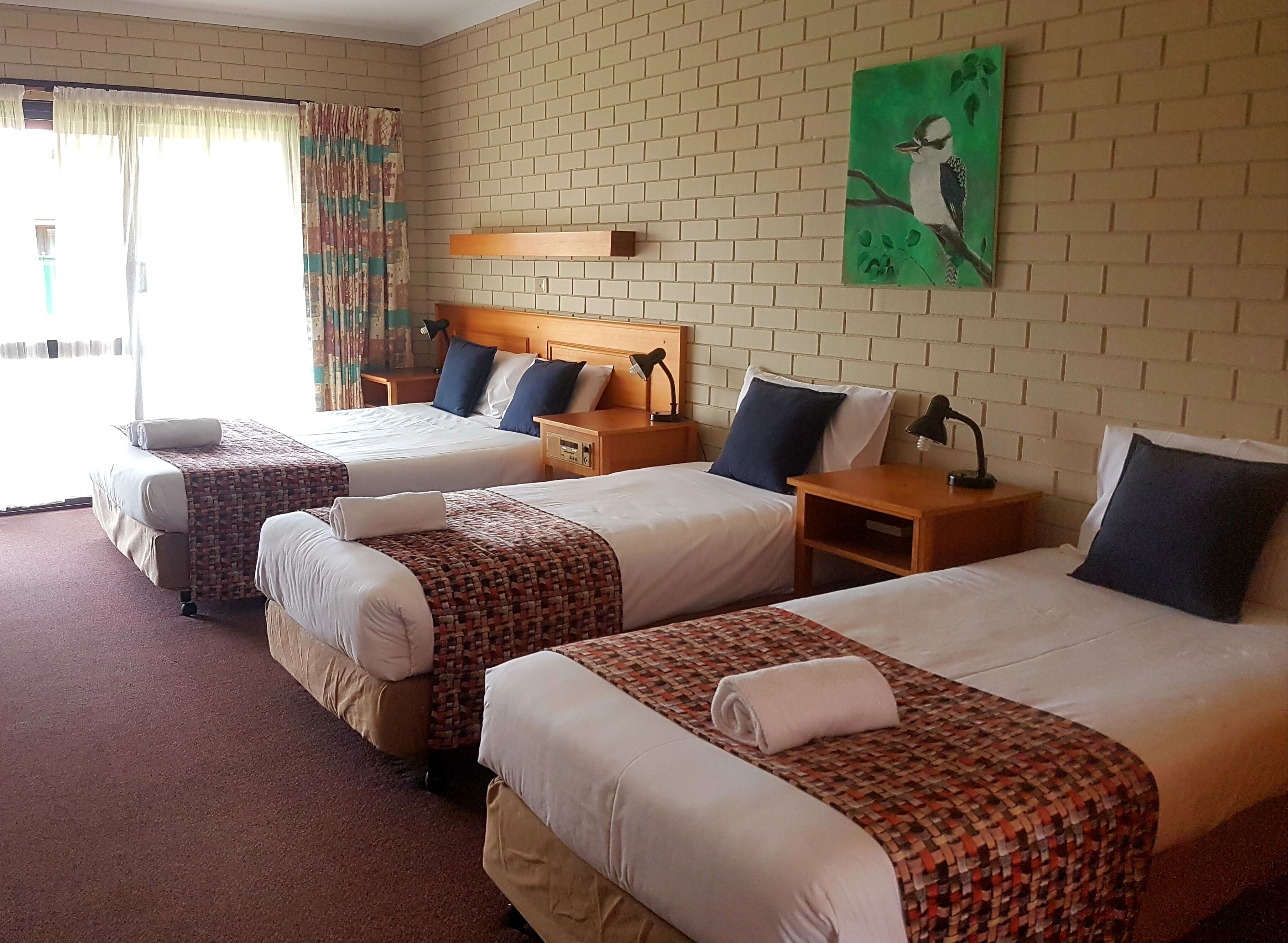 Habitación de hotel con tres camas, cada una con colcha y almohadas estampadas, cabeceras de madera y mesitas de noche con lámparas.