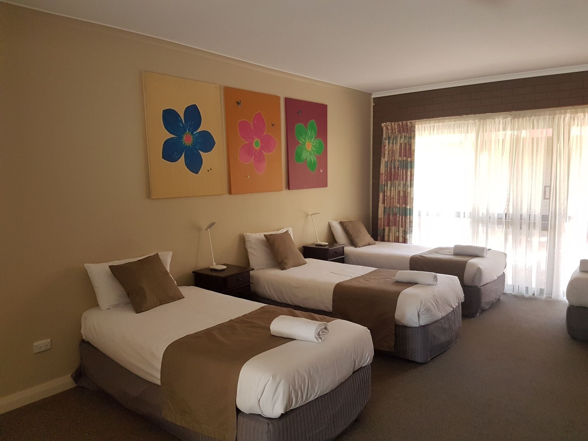 Ett hotellrum med tre sängar, ett stort fönster med tunna, genomskinliga gardiner och tre blomstermålningar på väggen.