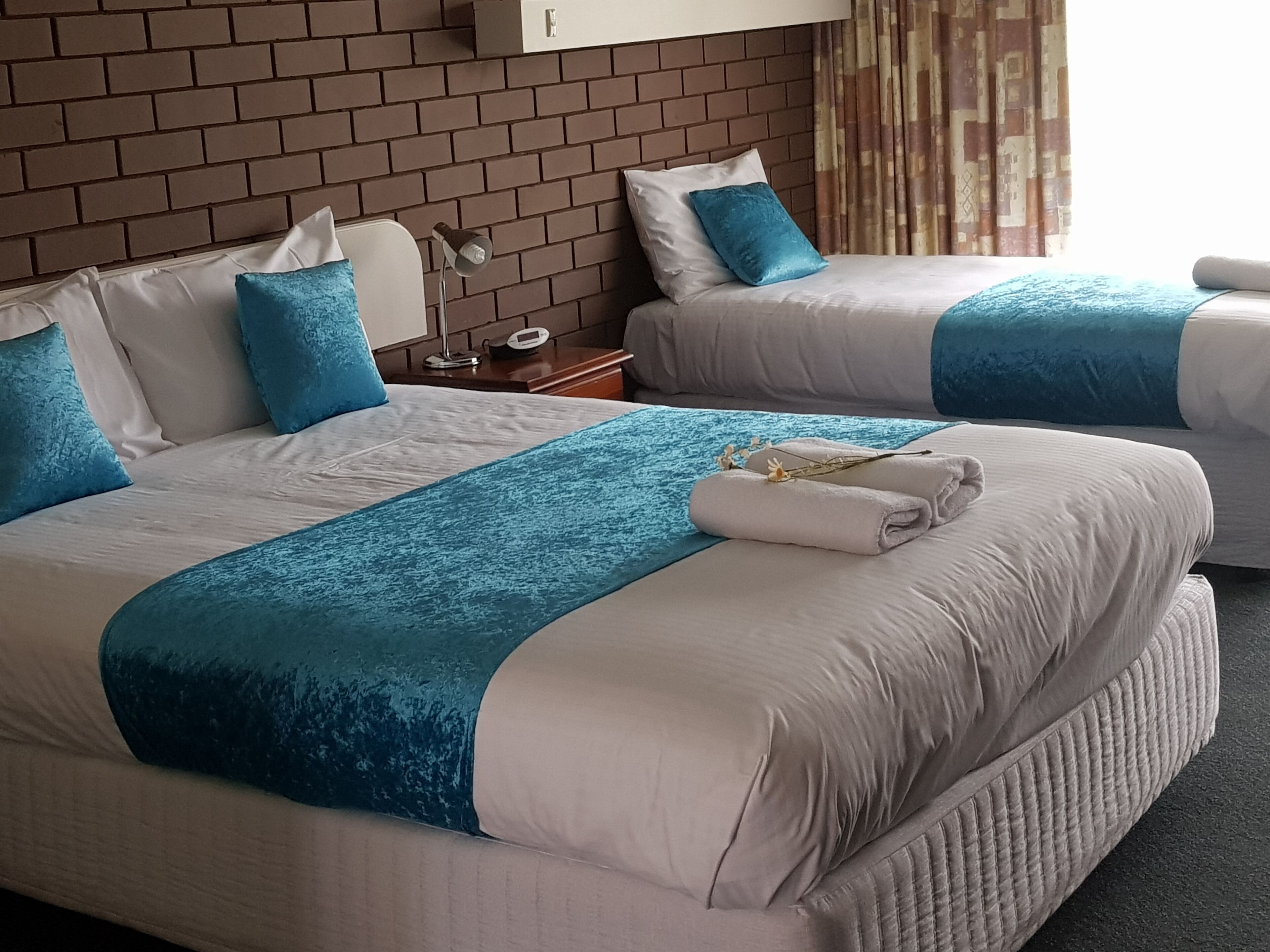 Hotellihuoneessa on kaksi sänkyä, joissa on valkoiset vuodevaatteet ja siniset yksityiskohdat. Sänkyjen välissä on yöpöytä, jossa on lamppu ja kello.