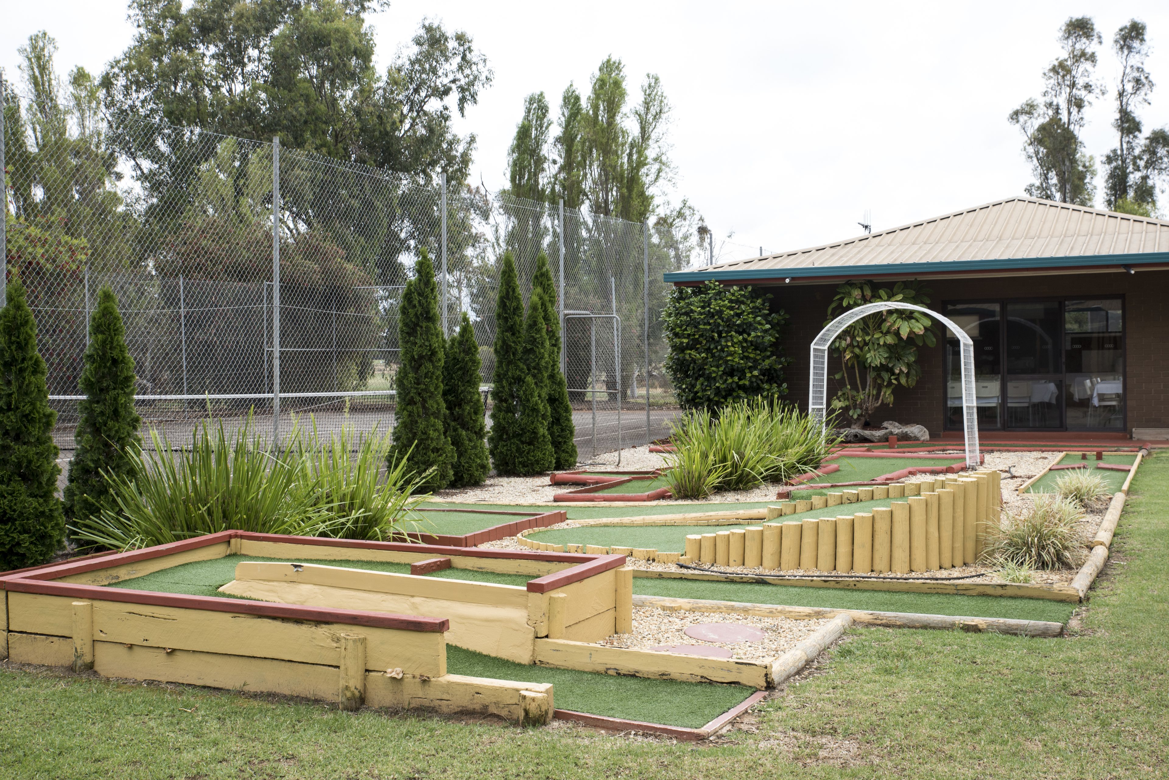 Un parcours de mini-golf doté d’une zone délimitée par une clôture, d’un gazon artificiel vert et d’un bâtiment avec un toit végétalisé en arrière-plan.