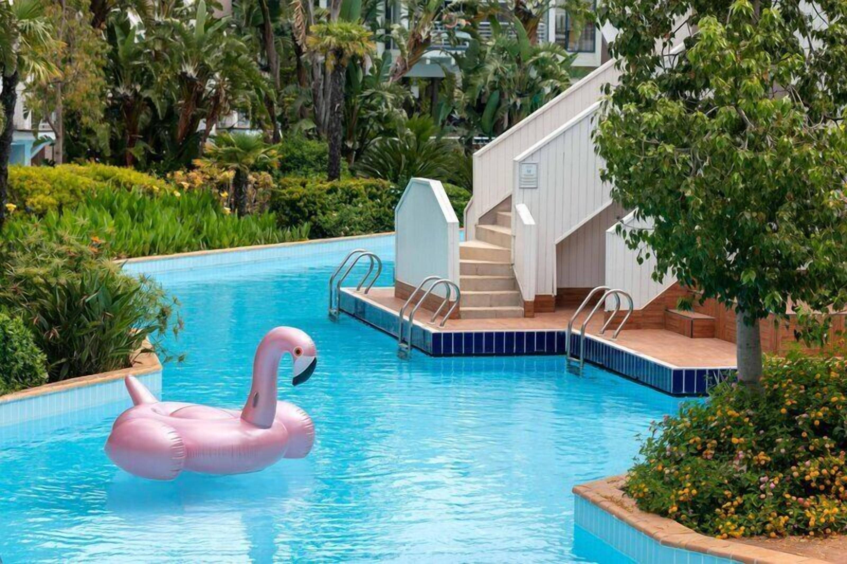 Een zwembad met een roze flamingo drijvend op het water, omringd door weelderige begroeiing en een wit gebouw met trappen.