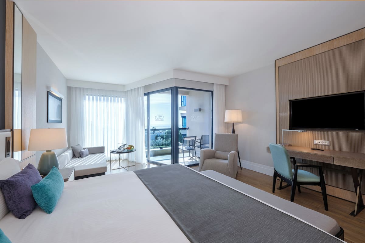 A modern hotel room with a large bed, a desk, a chair, a TV, and a balcony with a view.