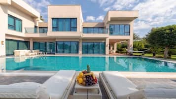 Ein modernes Haus mit zwei Etagen, einem Schwimmbecken, Liegestühlen und einer Obstschale.