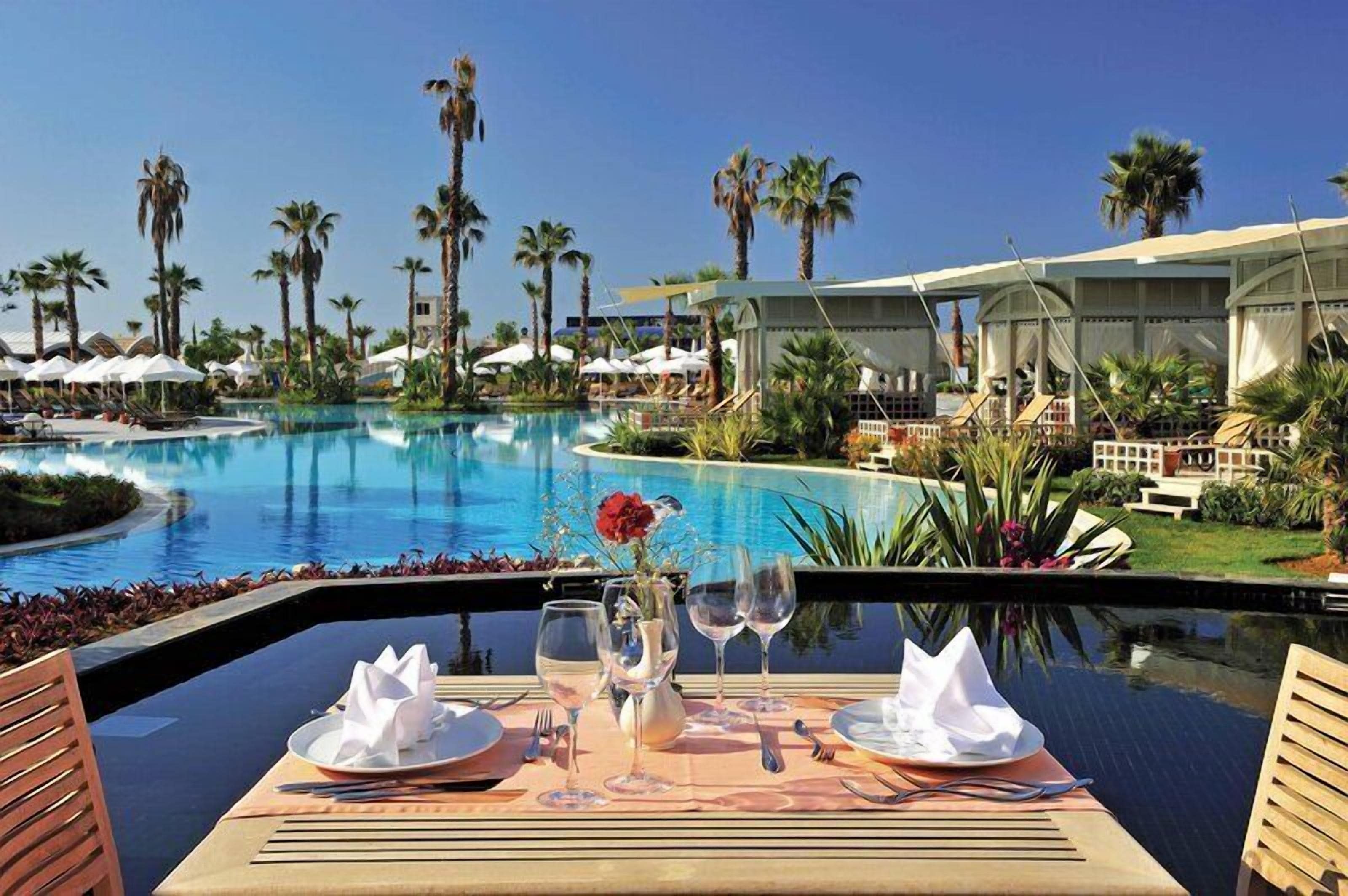 A dining table set with plates, glasses, and a vase with a flower, overlooking a pool with palm trees and a resort building.