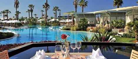 A dining table set with plates, glasses, and a vase with a flower, overlooking a pool with palm trees and a resort building.