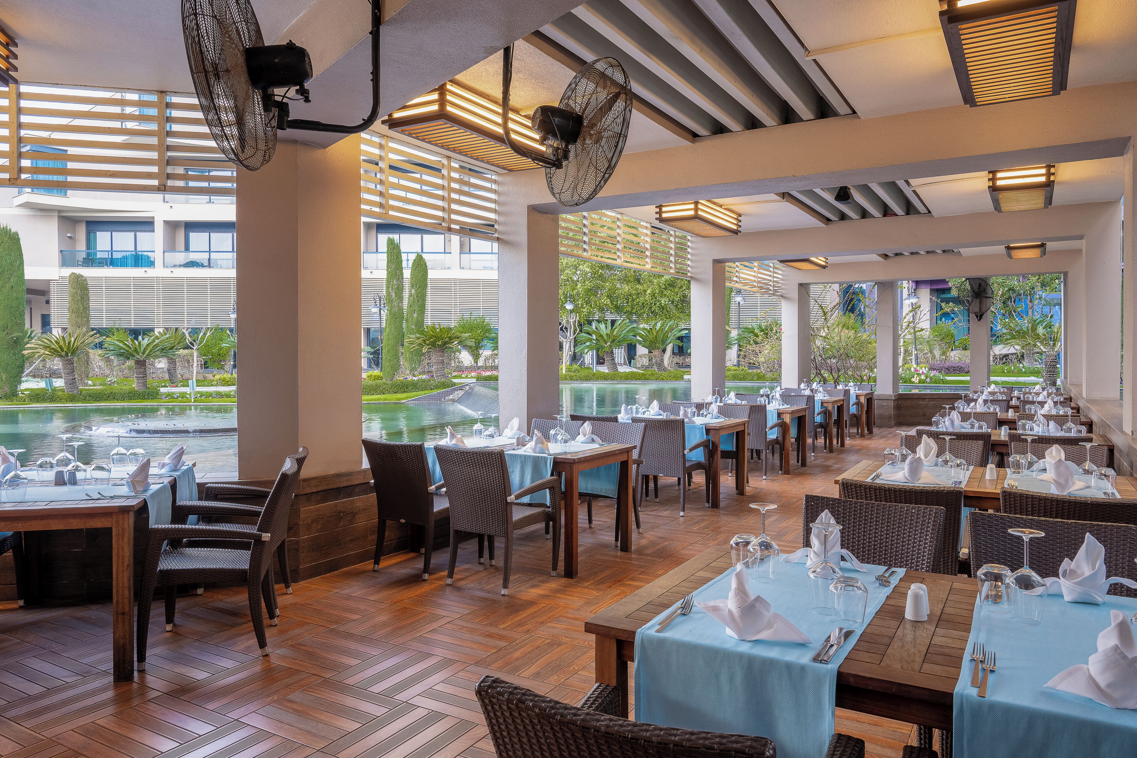 A restaurant with tables set for dining, large fans, and a view of a building and greenery.