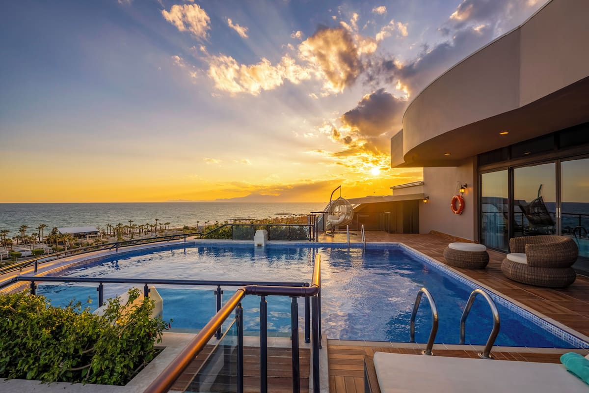 A pool with a view of the ocean at sunset, a fan, and outdoor seating.