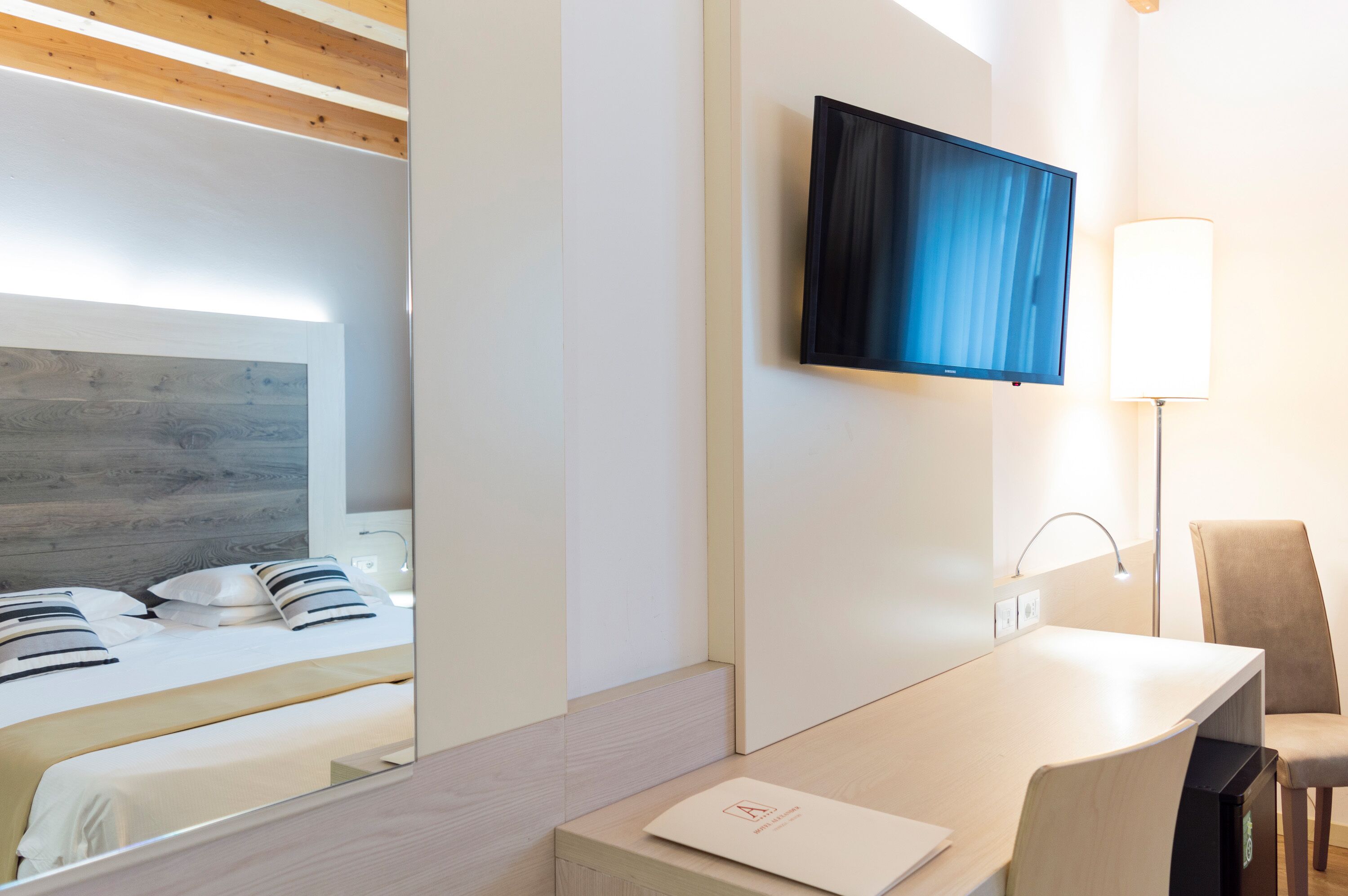 A modern hotel room with a bed, a desk, a chair, a TV mounted on the wall, and a lamp.