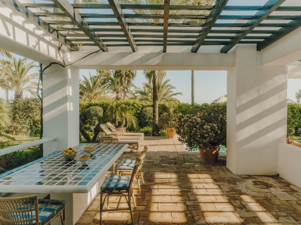 A covered outdoor area with a table and chairs, surrounded by greenery and a pool.